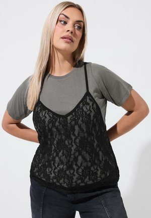 Kobieta z długimi blond włosami, ubrana w szarą koszulkę pod czarnym koronkowym topem i ciemne jeansy, patrząca na bok z rękami za plecami.