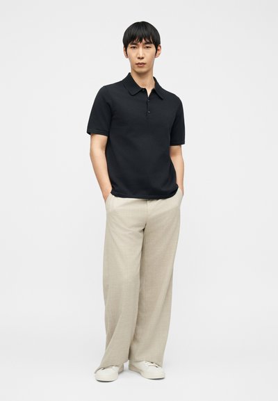 Άνδρας που στέκεται με τα χέρια στις τσέπες, φορώντας μαύρο πόλο μπλουζάκι, μπεζ παντελόνι με φαρδιά μπατζάκια και λευκά αθλητικά παπούτσια, μπροστά σε απλό λευκό φόντο.