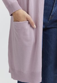 Cardigan en maille violet clair avec des poignets et un ourlet côtelés, présentant une poche à l'avant. Porté sur un jean en denim foncé. Texture lisse.