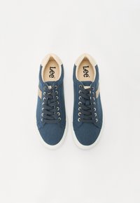 Blue canvas sneakers feature beige accents and brown laces, with a textured surface and a white rubber sole. Brand logo visible inside.