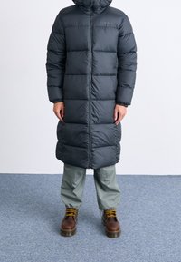 Lång, mörkgrå puffjacka med huva, med kviltade sektioner och dragkedja framtill. Kombinerad med ljusgröna byxor och bruna stövlar.