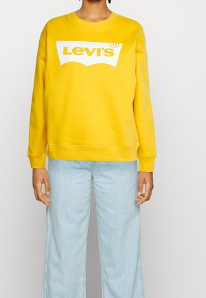 Osoba ubrana w jaskrawożółtą bluzę Levi's z dużym białym logo i jasnoniebieskie jeansy o szerokich nogawkach, stojąca na jednolitym tle.