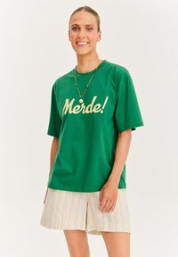 Zelené bavlnené tričko s potlačou "Merde!" v krémovej farbe. Voľný strih s krátkymi rukávmi. Kombinované s svetlo pruhovanými šortkami v pastelových odtieňoch.