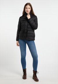 Svart vadderad jacka med hög krage, framslutning med dragkedja, dragsko i midjan och långa ärmar. Kombination med blå jeans och bruna snörkängor.