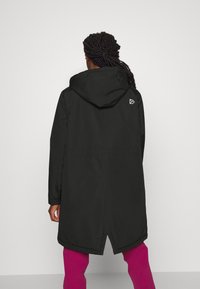 Zwarte, lang model parka-jas met capuchon, voorzien van een achterventilatie en een klein logo op de rechter schouder, gedragen over roze broek.