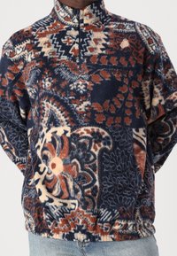 Pullover in pile con collo alto, caratterizzato da un motivo paisley in blu, arancione e crema. Chiusura con cerniera e tessuto morbido e strutturato.