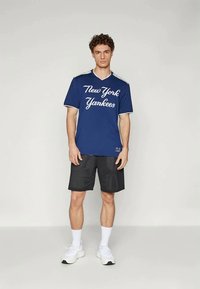 Camisola azul com o texto "New York Yankees" em branco, mangas curtas, decote em V, combinada com calções de malha pretos e sapatilhas brancas.