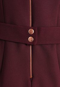 Burgundy jumpsuit med synlig dragkedja, två kopparknappar och ett brett bälte som dras åt i midjan, tillverkat av texturerat tyg.