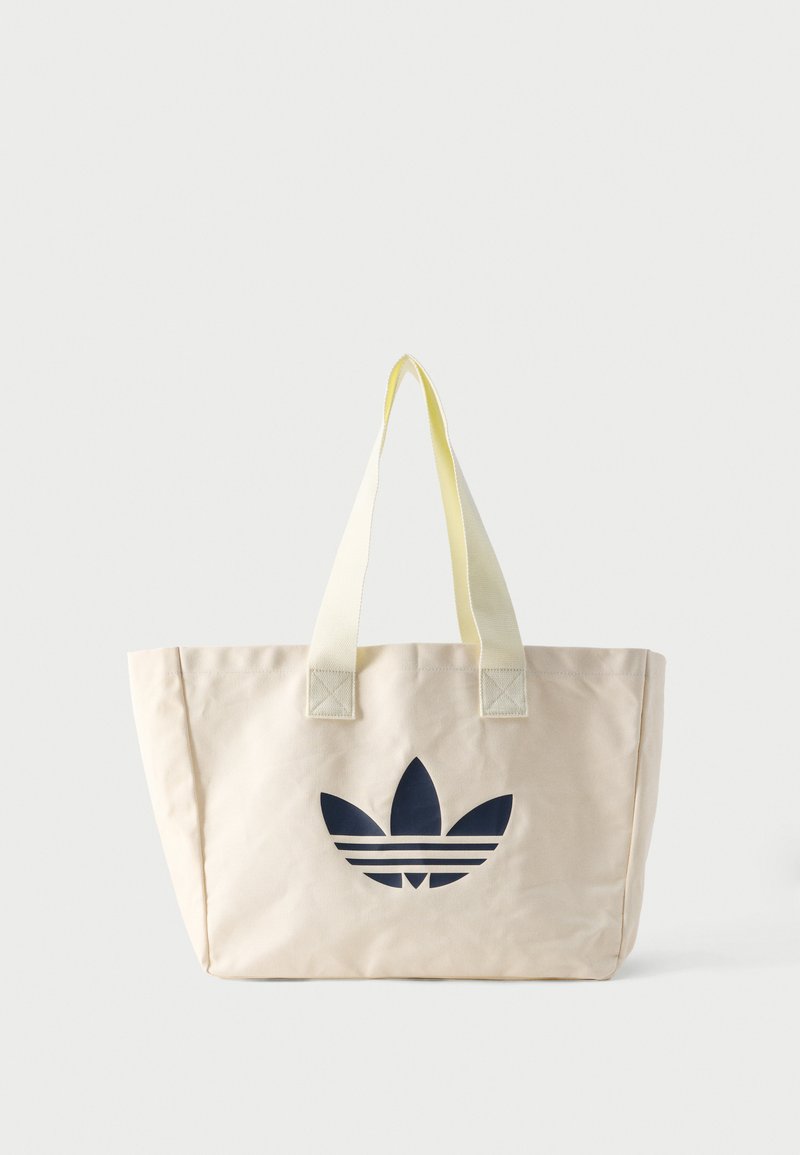 Béžová plátená taška cez rameno s bledozltými rukoväťami a veľkým čiernym logom Adidas trefoil uprostred prednej strany.