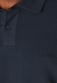 Detailní záběr na tmavě modrou pletenou polo košili s límečkem, knoflíkovou légou a vyšitým logem "BOSS" na hrudi.