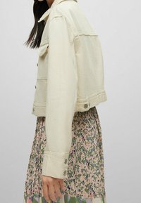 Krémová denimová bunda s širokým střihom, velkým límcem, zapínáním na knoflíky a dvěma předními kapsami, nošená přes pastelovou květinovou sukni.