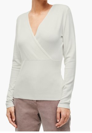 Vrouw draagt een wit, langarmig omslagvest gecombineerd met lichtbruine broek, staand tegen een effen witte achtergrond.