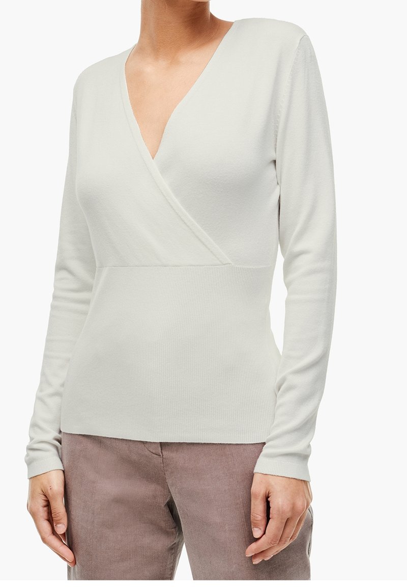 Femme portant un pull blanc à manches longues de style cache-cœur assorti à un pantalon marron clair, debout devant un fond blanc uni.