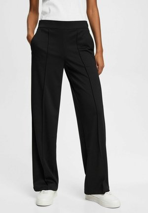 Personne portant un pantalon noir à jambes larges avec des plis devant, mains dans les poches, assorti à des baskets blanches et un haut blanc sans manches.