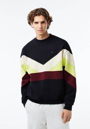 Sudadera azul marino con patrón de chevron en crema, lima y burdeos. Presenta un cuello redondo y puños elásticos. Logo de Lacoste en el pecho.
