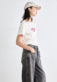 Camiseta blanca de manga corta con texto rojo, combinada con pantalones de mezclilla grises de cintura alta. El modelo lleva una gorra de béisbol en color beige claro y tiene las manos en los bolsillos.