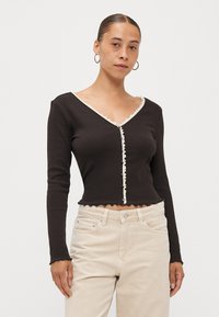 Black long-sleeve top with V-neck, contrast scalloped trim, and front buttons. Cropped design with a textured finish, paired with light pants.