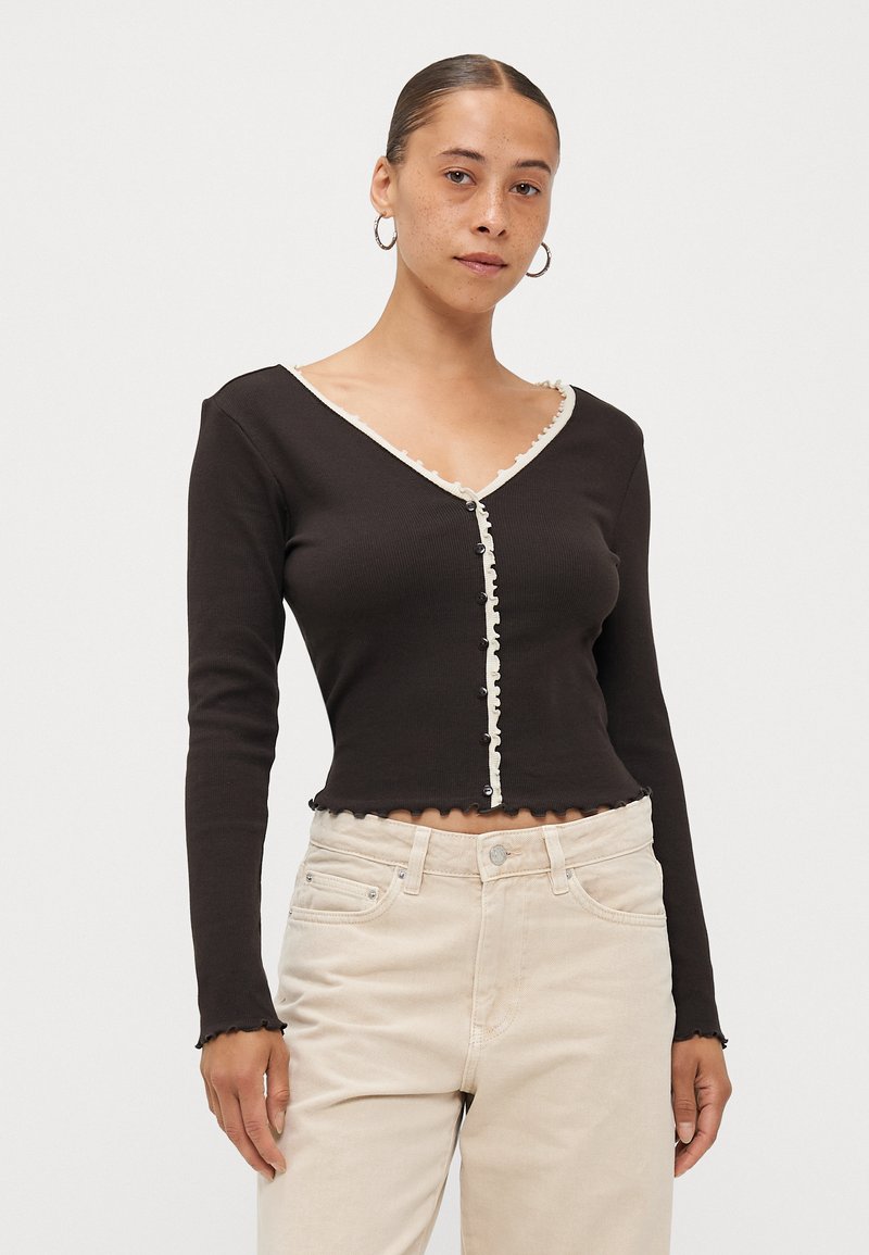 Black long-sleeve top with V-neck, contrast scalloped trim, and front buttons. Cropped design with a textured finish, paired with light pants.