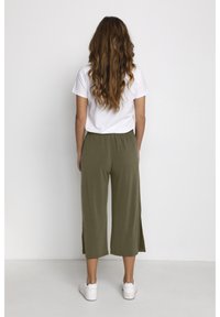 Olivgrön vida byxor med sidoklackar, parat med en vit kortärmad skjorta och vita sneakers. Bakgrunden är helt vit.