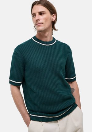Jeune homme aux cheveux mi-longs portant un pull en maille vert foncé à manches courtes avec bordure rayée blanche et un pantalon clair, mains dans les poches.