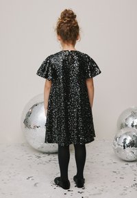 Schwarzes Paillettenkleid mit kurzen Puffärmeln, knielang und mit Schlüsselloch-Detail am Rücken. Schwarze Strumpfhosen und Schuhe vervollständigen das Outfit. In der Nähe sind Discokugeln zu sehen.