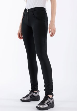 Leggings neri con una texture liscia, dotati di un alto punto vita, due tasche frontali e una chiusura a due bottoni. Abbinati a scarpe sportive con accenti argentati.