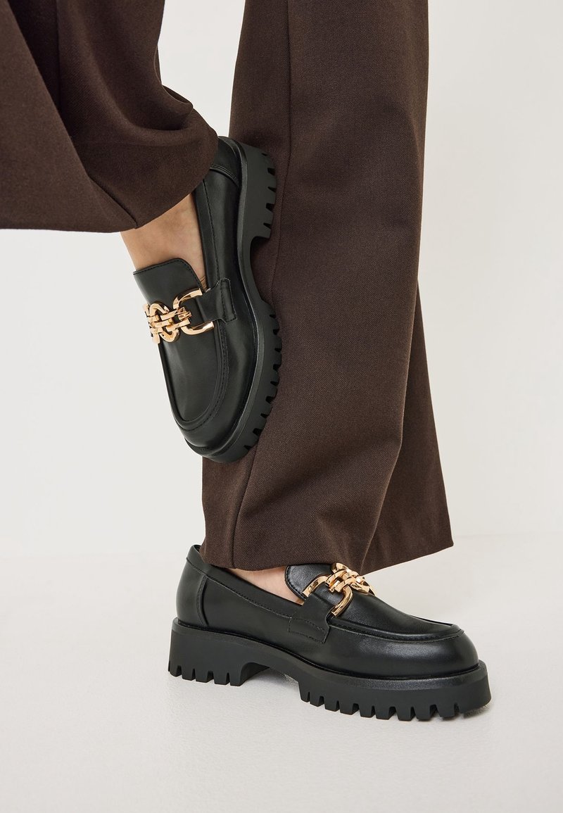 Mocassins noirs épais avec un détail chaîne dorée portés avec un pantalon marron à jambes larges sur un fond neutre.