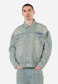 Svetlo modra jeans jakna s sproščenim krojem, vidnim šivom, sprednjimi žepi in gumbi na pritisak; ima teksturirano površino in ni podložena.