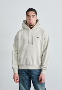 Felpa beige chiaro con cappuccio e lacci, tasca frontale e logo nero. Il tessuto sembra morbido, con polsini e orlo a coste. Portata con jeans blu.