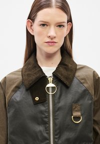 Dark green and black jacket with a corduroy collar, gold zipper, and ring hardware. Design features a textured finish and button details.