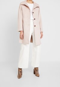 Manteau rose clair en mélange de laine avec col pointu, trois boutons et fentes latérales, associé à un pantalon large blanc et des bottines imprimé peau de serpent.