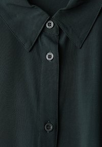 Mörkgrön skjorta med krage i slätt tyg, två svarta knappar och synliga sömnadsdetaljer längs kanterna på kragen.