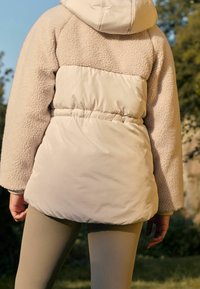 Beige hoodie-jack met een mix van pluizige fleece en gladde stof, met een getailleerde taille en geribde manchetten. Gecombineerd met strakke leggings.