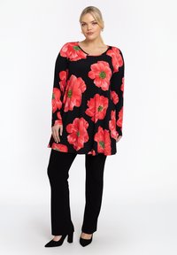 Haut à manches longues noir avec un grand motif floral rouge et des centres verts, associé à un pantalon droit noir et des talons assortis.