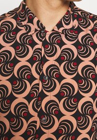 Chemise à manches courtes à boutons, présentant un motif circulaire noir et pêche avec des accents rouges, confectionnée dans un tissu léger à la texture lisse.