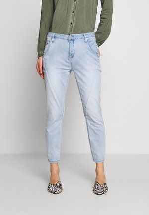 Osoba ubrana w jasnoniebieskie, zwężane dżinsy, oliwkową koszulę na guziki oraz czarno-białe, wzorzyste płaskie buty, stojąca na jednolitym tle.