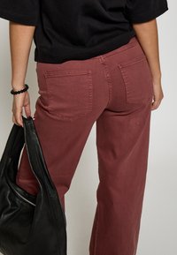 Kastanis plaša kāju bikses, izgatavotas no kokvilnas, ar aizmugures kabatām. Modelis tur melnu ādas somu un valkā īsu melnu topu.