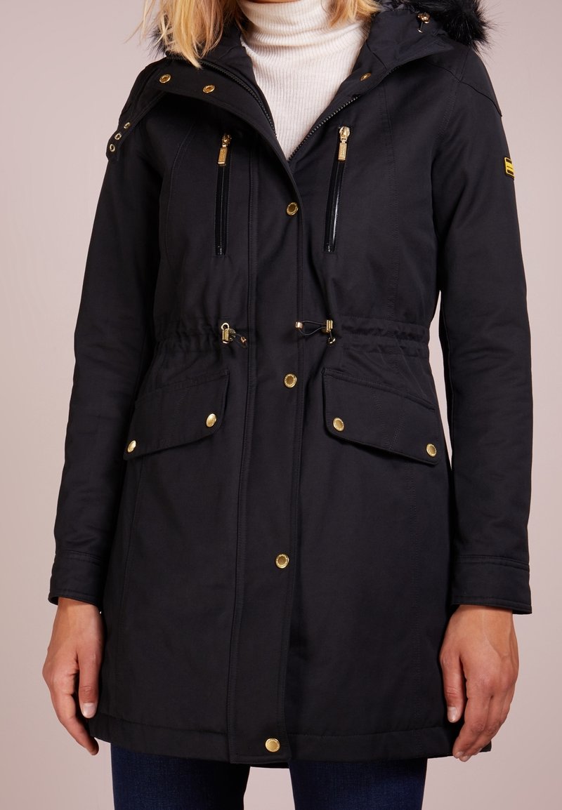 Schwarze Parkajacke mit futterbesetzter Kapuze, Taillenzug, goldfarbenen Beschlägen, zwei Fronttaschen und Reißverschlusstaschen an der Brust.