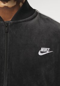 Svart bomberjacka med en slät, sammetsliknande yta. Har en framzipper, ribbad krage och en vit broderad Nike-logga på bröstet.