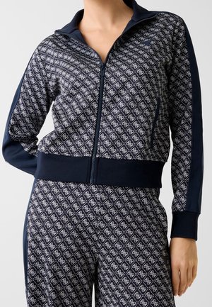 Femme portant une veste zippée bleu marine et un pantalon assorti avec un motif géométrique blanc et des panneaux latéraux bleu marine unis.