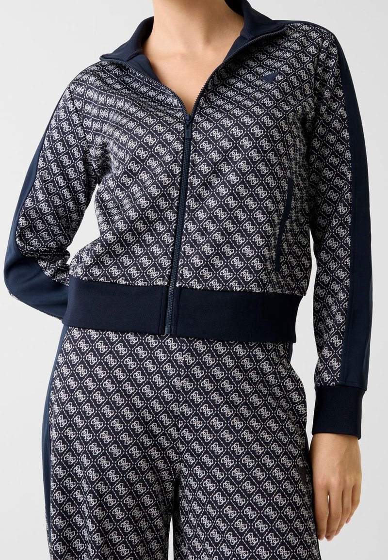 Femme portant une veste zippée bleu marine et un pantalon assorti avec un motif géométrique blanc et des panneaux latéraux bleu marine unis.