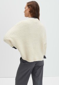 Donna con capelli castani che indossa un maglione bianco grosso e ampio e pantaloni grigio scuro, in piedi con la schiena e la testa rivolte a sinistra.