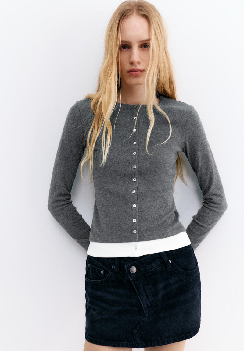 Fiatal nő hosszú szőke hajjal, szürke, gombos kardigánt visel fehér felső felett, és rövid fekete farmer szoknyát.