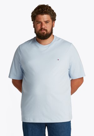 Mann mit Bart, der ein hellblaues kurzärmeliges T-Shirt und dunkelblaue Jeans trägt, steht mit den Händen hinter dem Rücken vor einem einfarbigen Hintergrund.