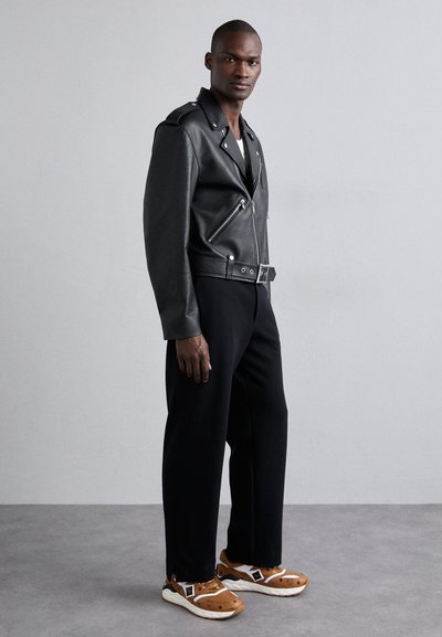 Veste de biker en cuir noir avec des fermetures éclair argentées, associée à un pantalon noir ample et des baskets marron avec des accents blancs et un motif texturé.