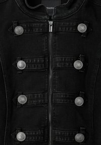 Veste noire avec une fermeture éclair centrale et six boutons décoratifs en métal sur des bandes de tissu horizontales en deux colonnes verticales.