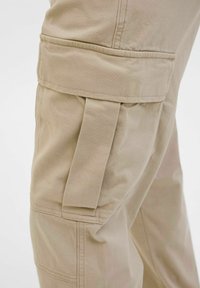 Pantalon cargo beige avec une grande poche latérale à rabat et un détail de sangle en tissu sur la cuisse.