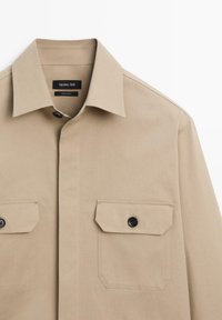 Camicia beige realizzata in tessuto liscio con colletto a punta, due tasche sul petto e bottoni neri. Linee pulite e design strutturato.