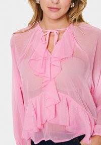 Blouse en voile rose avec des détails froncés, dotée d'une fermeture à nouer au cou et d'un ourlet superposé. Tissu doux et léger.