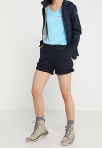 Námořnické outdoorové šortky v kombinaci s světle modrým tričkem s výstřihem do V a tmavou bundou, doplněné šedými turistickými botami a texturovanými šedými ponožkami.